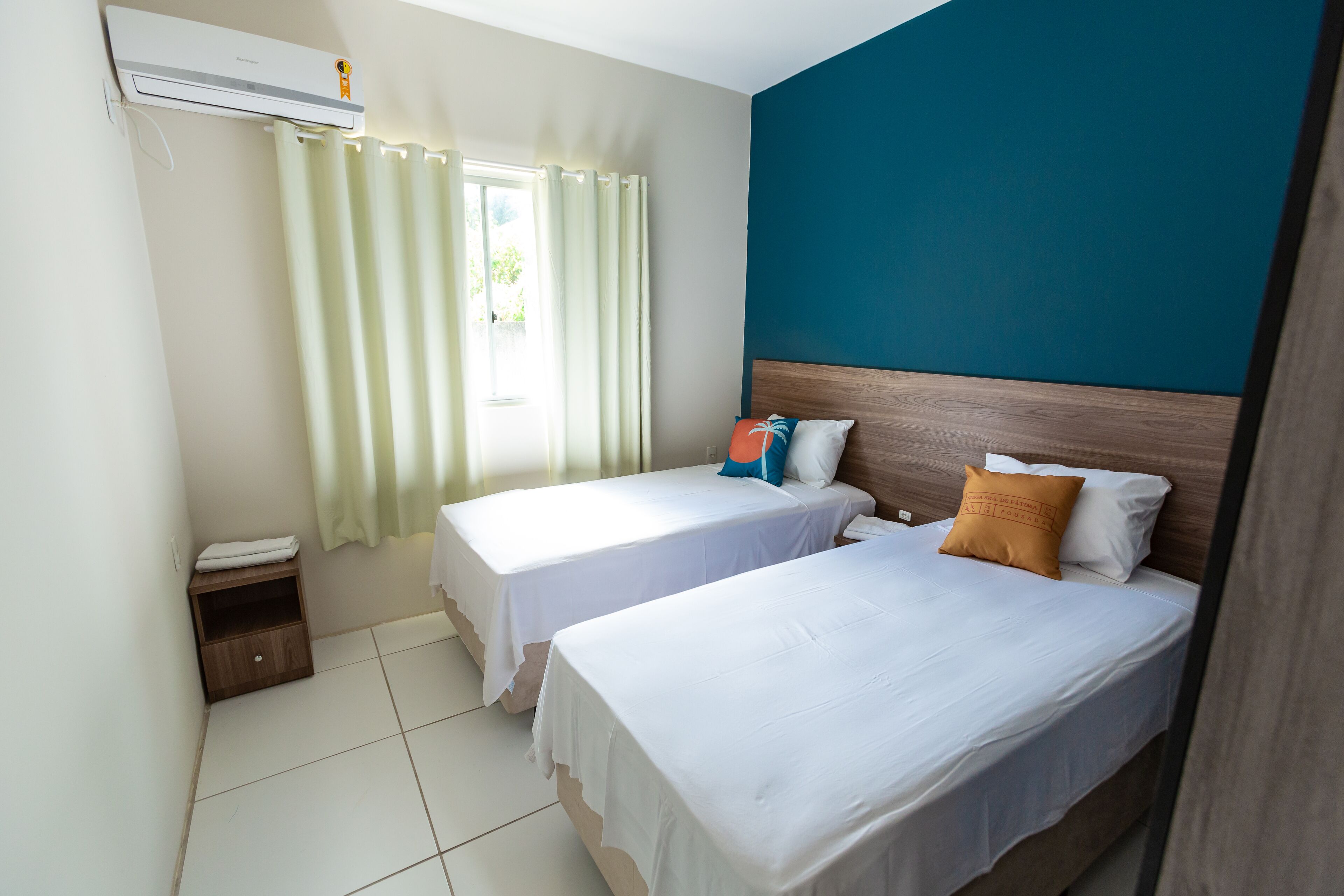 Habitación de hotel con dos camas, una ventana con cortinas, cabecera de madera y una mesita de noche de madera.