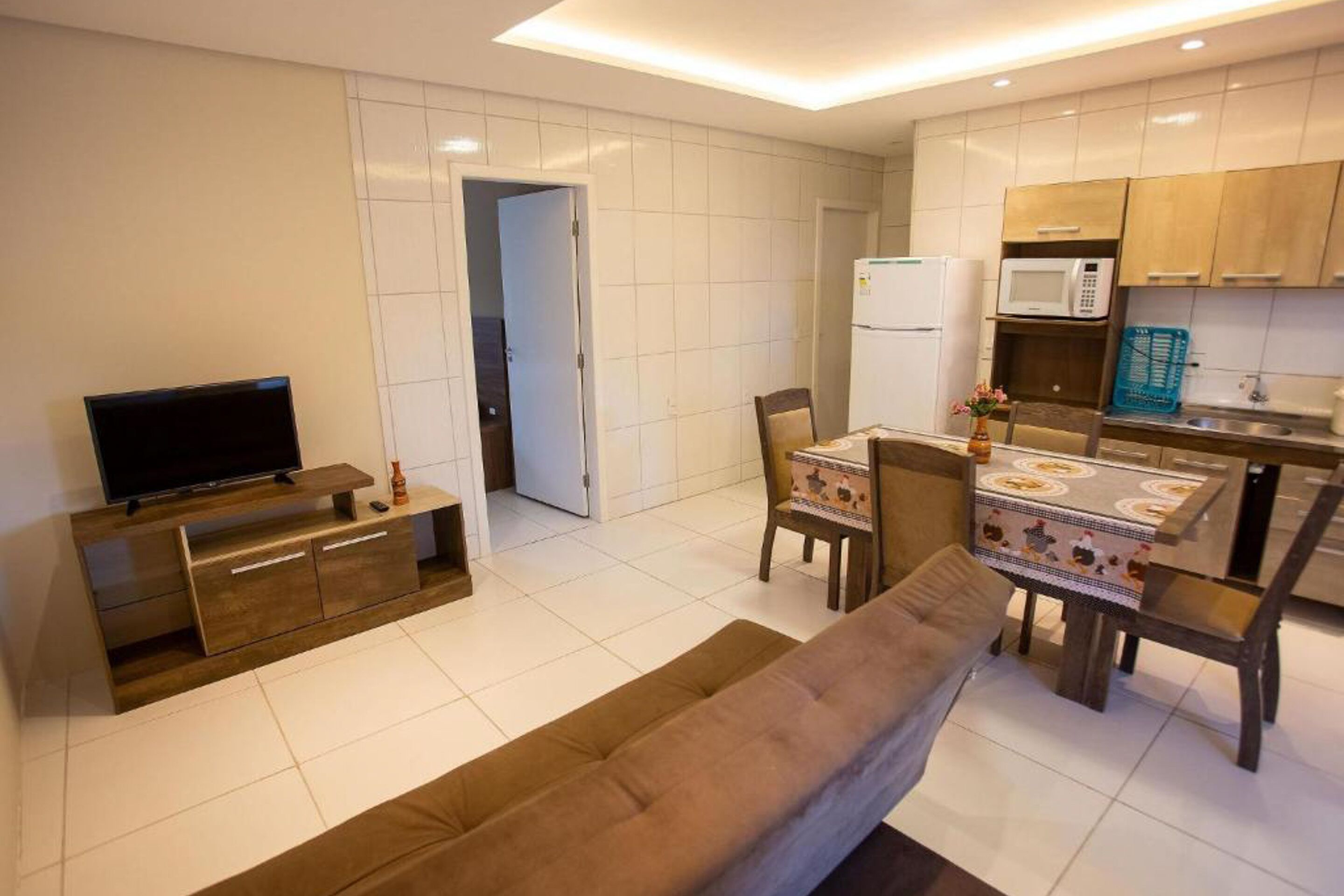 A modern kitchen with white tiles, a dining table with chairs, a sofa, and a TV on a wooden stand.