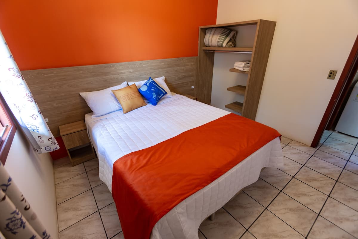 Un lit avec une couvre-lit blanche matelassée et une couverture orange, une tête de lit en bois et une étagère en bois où sont pliées des serviettes.