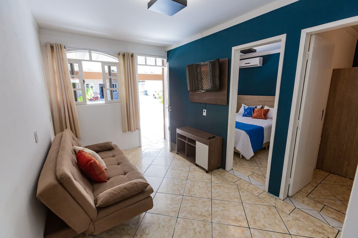 Une pièce comprenant un canapé, un lit recouvert d’une couverture bleue, un téléviseur fixé au mur et une porte donnant sur une autre pièce.