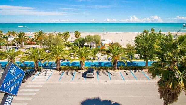 A parking lot with a sign for Excelsior Hotel, palm trees, and a beach in the background. - Hotel Excelsior (Alba Adriatica)