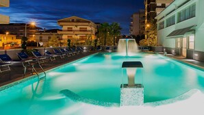 A swimming pool with a water feature in the center, surrounded by lounge chairs and a building with balconies. - Hotel Excelsior (Alba Adriatica)