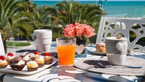 A table with pastries, a glass of orange juice, and a heart-shaped mug, set against a backdrop of a beach and palm trees. - Hotel Excelsior (Alba Adriatica)