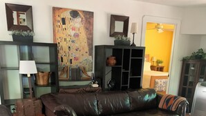 Una sala de estar con un sofá de cuero marrón, un cofre de madera y un tapiz colorido. Hay un cuadro en la pared y un aparador con un espejo.