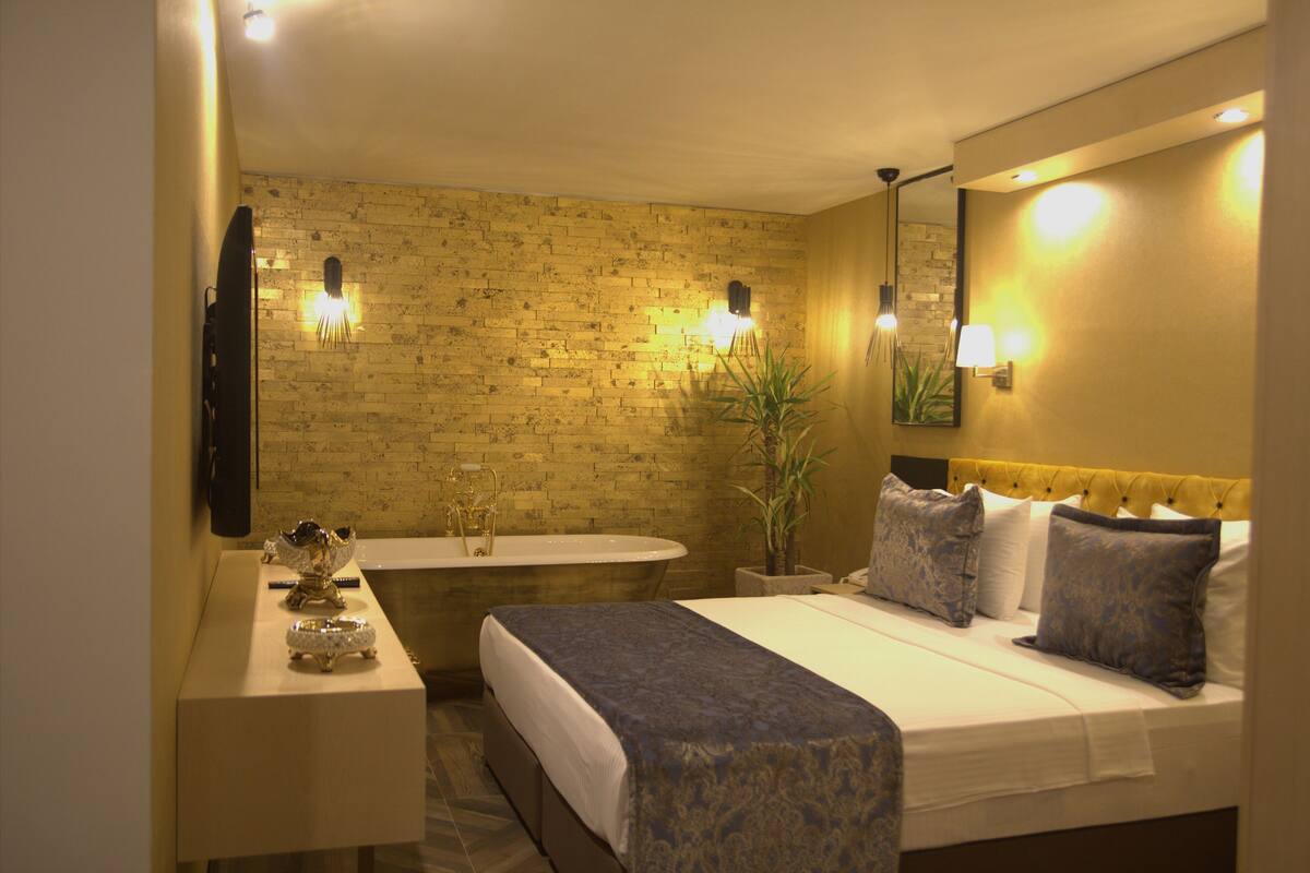 Hotelkamer met een bakstenen muur, groot bed met blauw beddengoed, nachtkastje met decoratieve schaal, badkuip en wandlampen.
