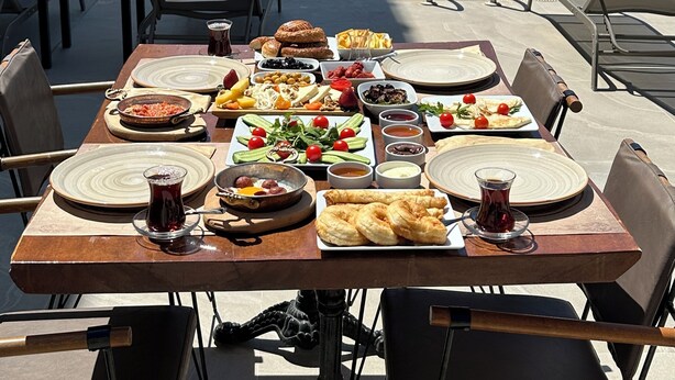 Une table à manger dressée avec différents plats et boissons, donnant sur une piscine bordée de chaises longues et d’ombrelles.
