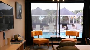 Une chambre d’hôtel avec un lit, un bureau avec une télévision, une chaise, une table avec un verre de vin, et une vue sur une piscine et des parasols à l’extérieur.