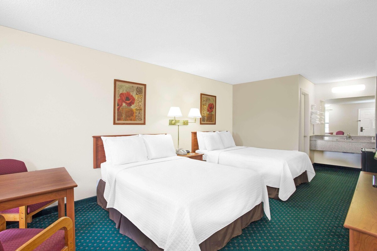 A hotel room with two beds, a desk, a chair, a nightstand with lamps, and a bathroom visible through an open door.