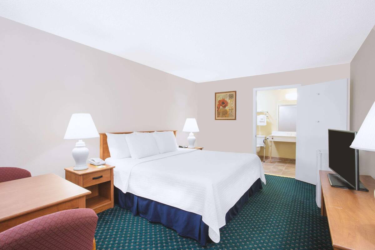 A hotel room with a bed, bedside tables, a desk, a chair, a television, and a bathroom visible through an open door.