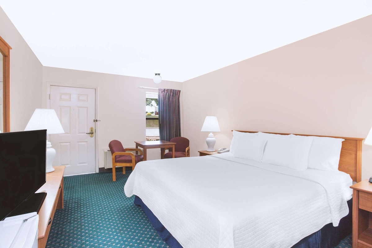 A hotel room with a bed, a desk with a TV, two chairs, a table, a lamp, and a window with curtains.