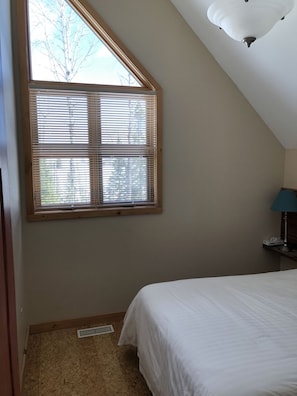 Un dormitorio con techo inclinado, una ventana con persianas, una cama y una mesita de noche con lámpara.