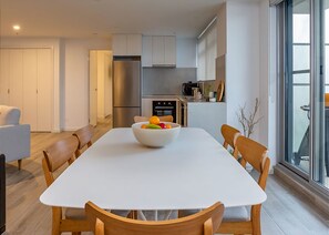 A modern kitchen with a stainless steel refrigerator, a dining table with a bowl of fruit, and wooden chairs.