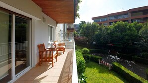 Ein Balkon mit Holzkufen und Tisch, ein Fliesenboden und Blick auf einen Garten mit einer Bank sowie ein Gebäude im Hintergrund.