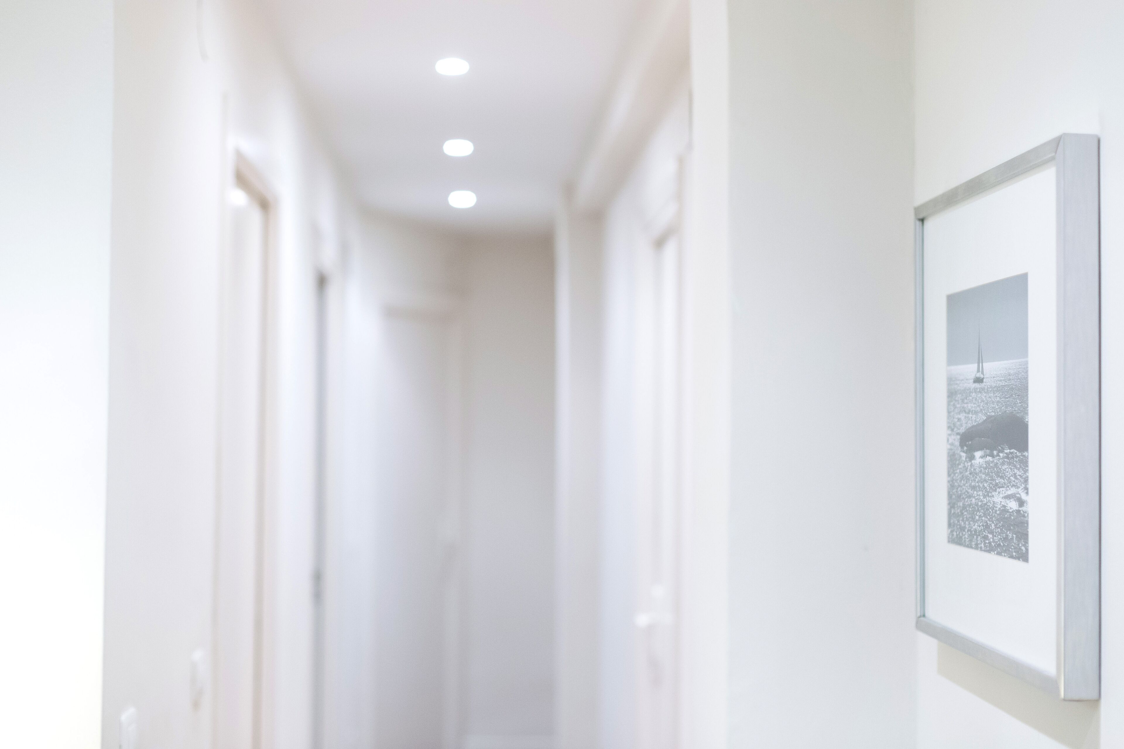 A narrow, well-lit hallway with white walls and doors, leading to a room with a framed picture on the wall.