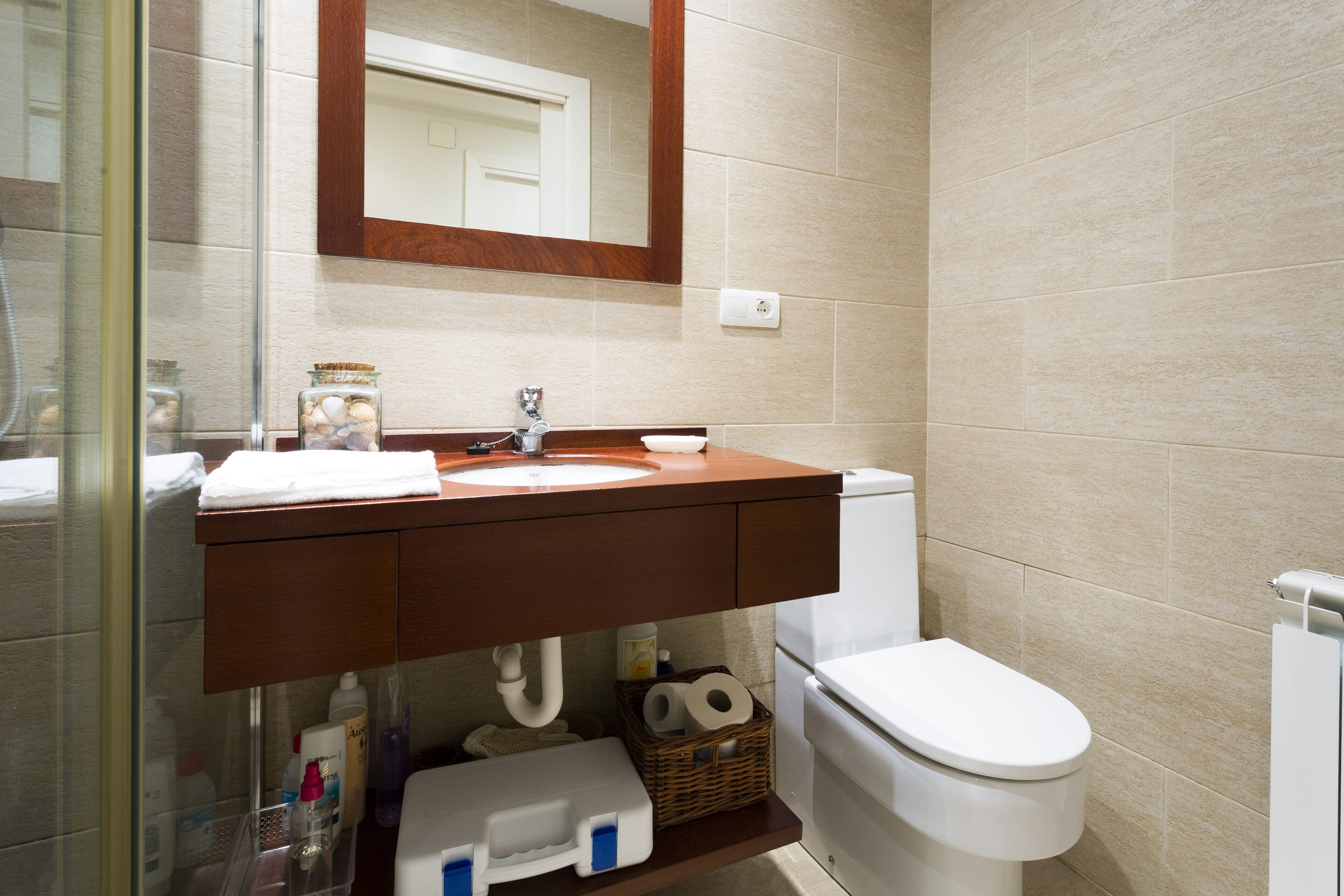 A bathroom with a wooden vanity, white sink, and toilet. There is a mirror above the sink and a glass shower enclosure to the left.