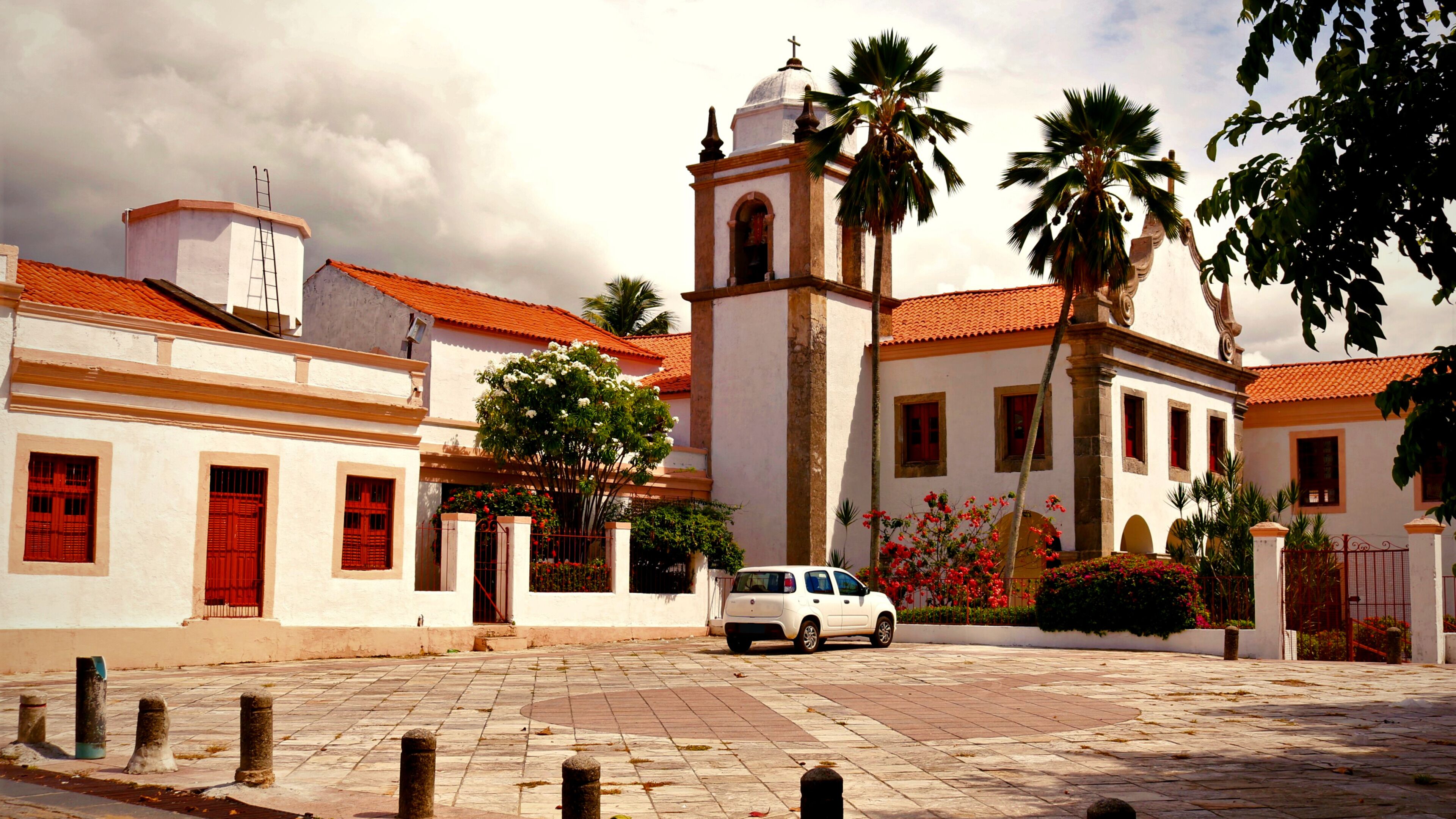 Un edificio blanco con puerta roja y un campanario, rodeado de palmeras y un patio empedrado.