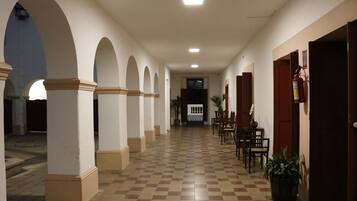 Un pasillo con suelo a cuadros, paredes blancas y arcos. Se observan puertas a la derecha y un extintor de incendios en la pared.