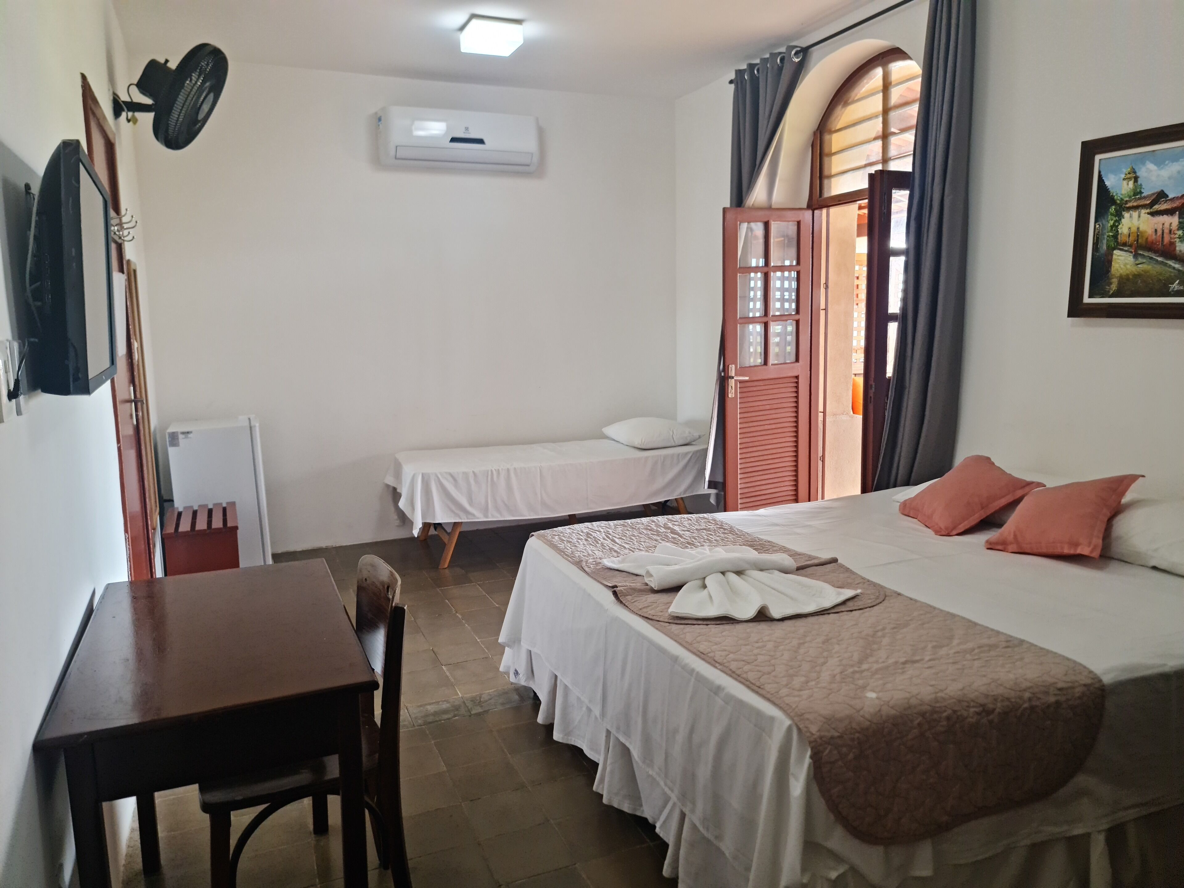 A hotel room with a bed, a desk, a chair, a small table, an air conditioner, a window with curtains, and a painting on the wall.