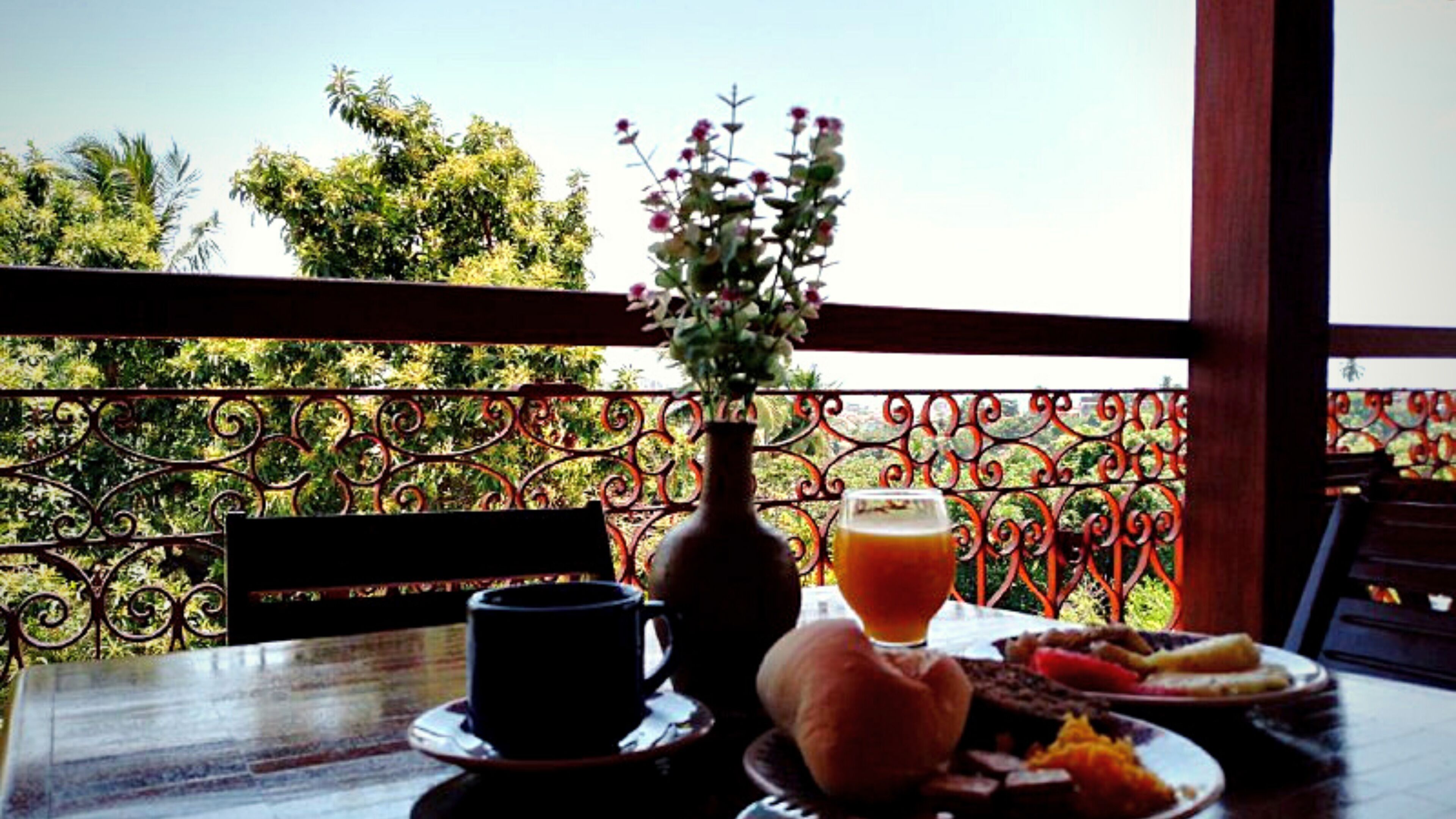 A dining table with a vase of flowers, a cup, and a glass of orange juice, set against a balcony railing with a view of trees.