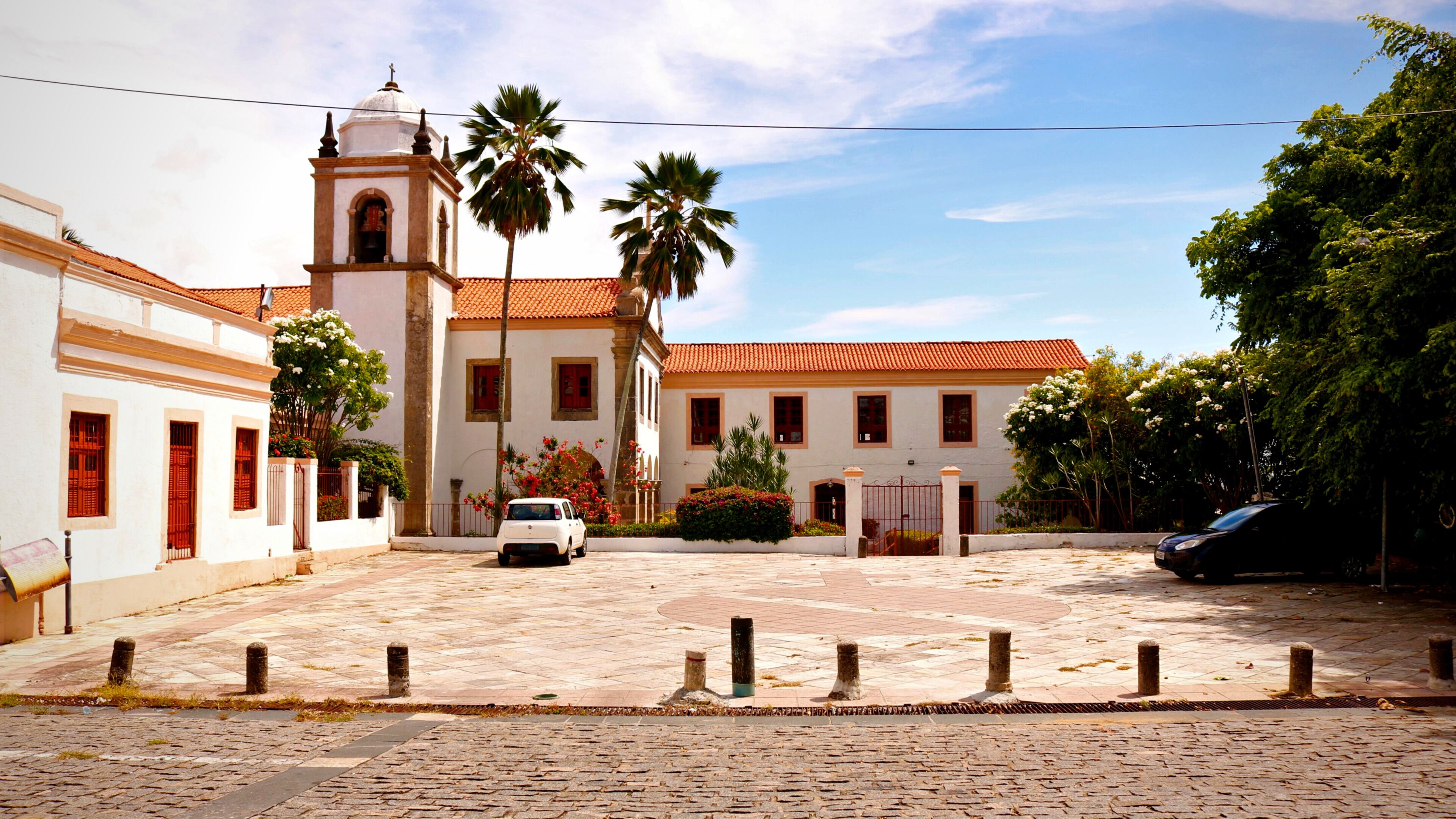 A courtyard with a white building featuring a bell tower, palm trees, and a paved area with bollards.