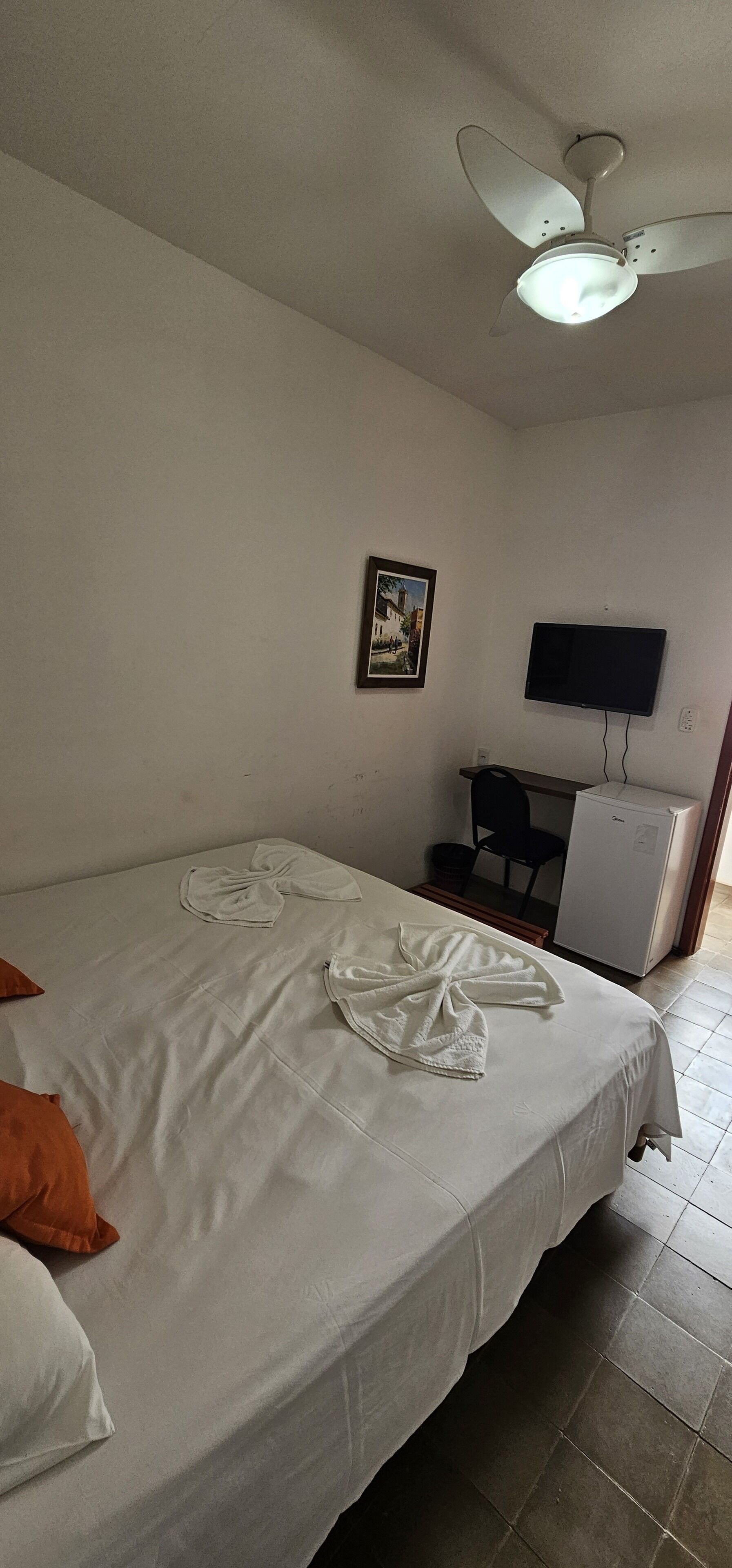 Quarto de hotel com cama, ventoinha no teto, televisão na parede e um pequeno frigorífico.