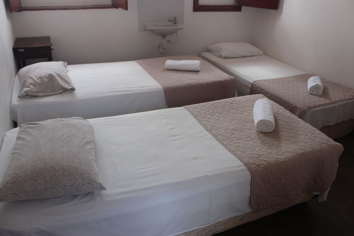 A hotel room with two single beds, a small bedside table, a wall-mounted sink, and a window with wooden frames.