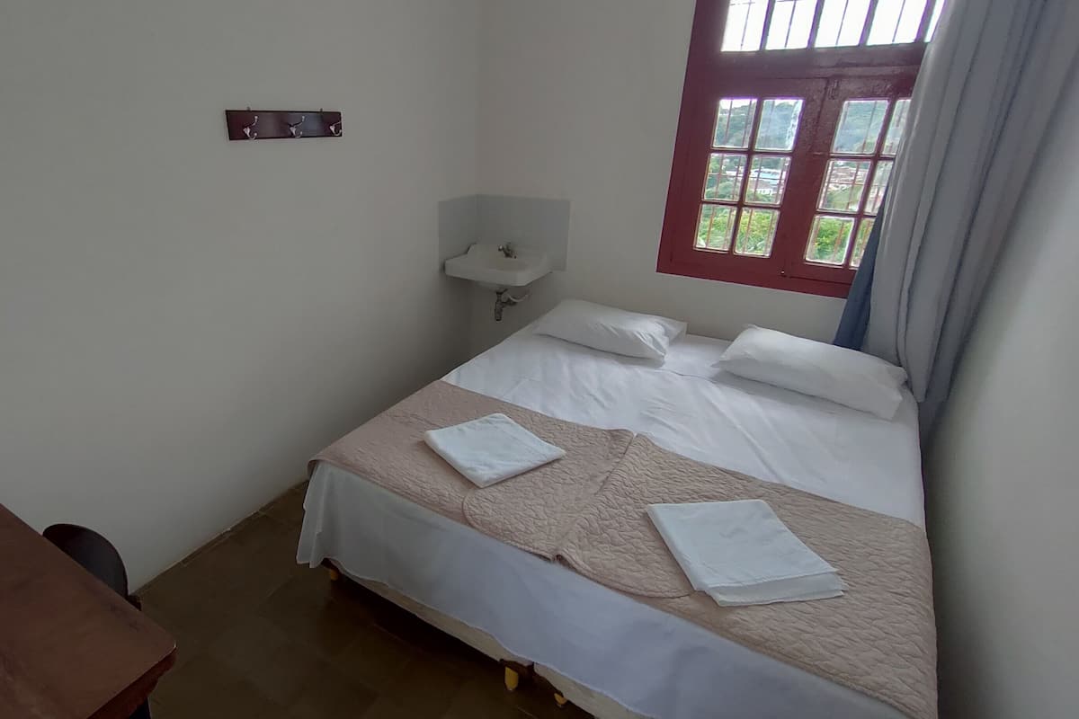 A small, clean room with a single bed, a bedside table, a window with curtains, and a wall-mounted sink.