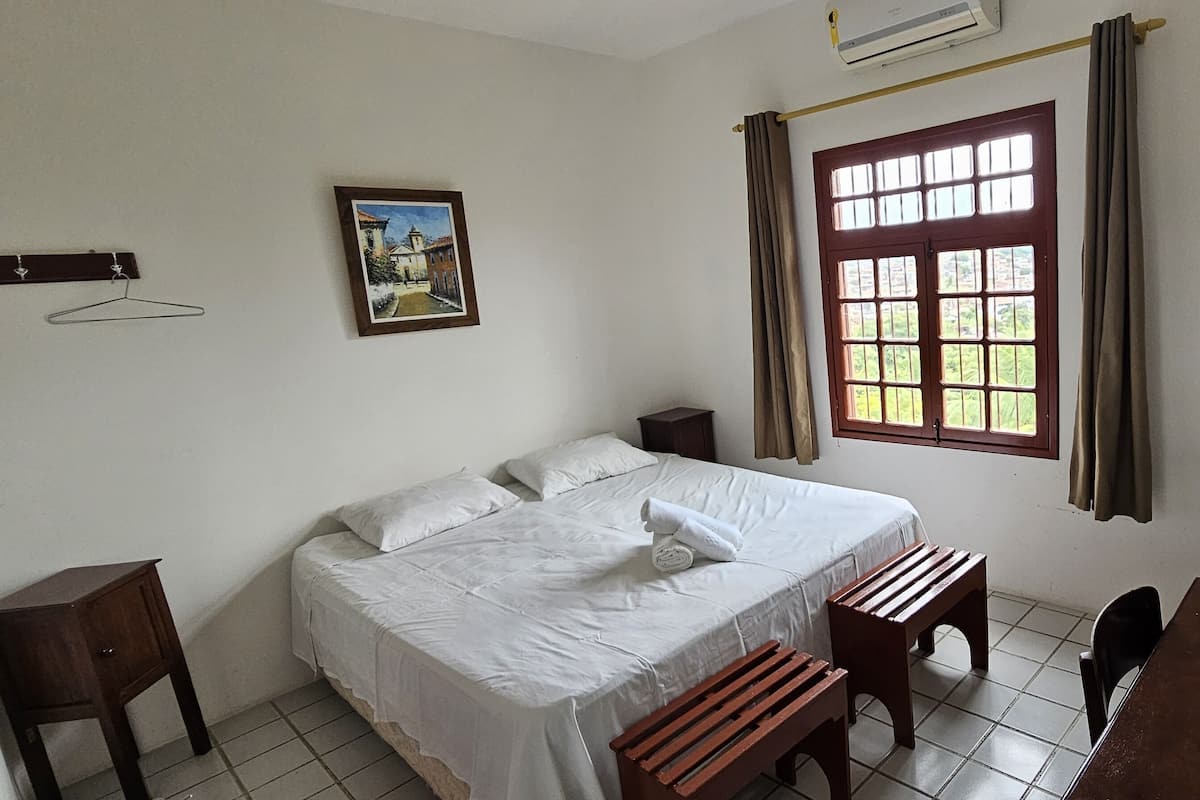A bedroom with a bed, bedside tables, a desk, a chair, a hanging coat rack, a picture on the wall, and a window with curtains.