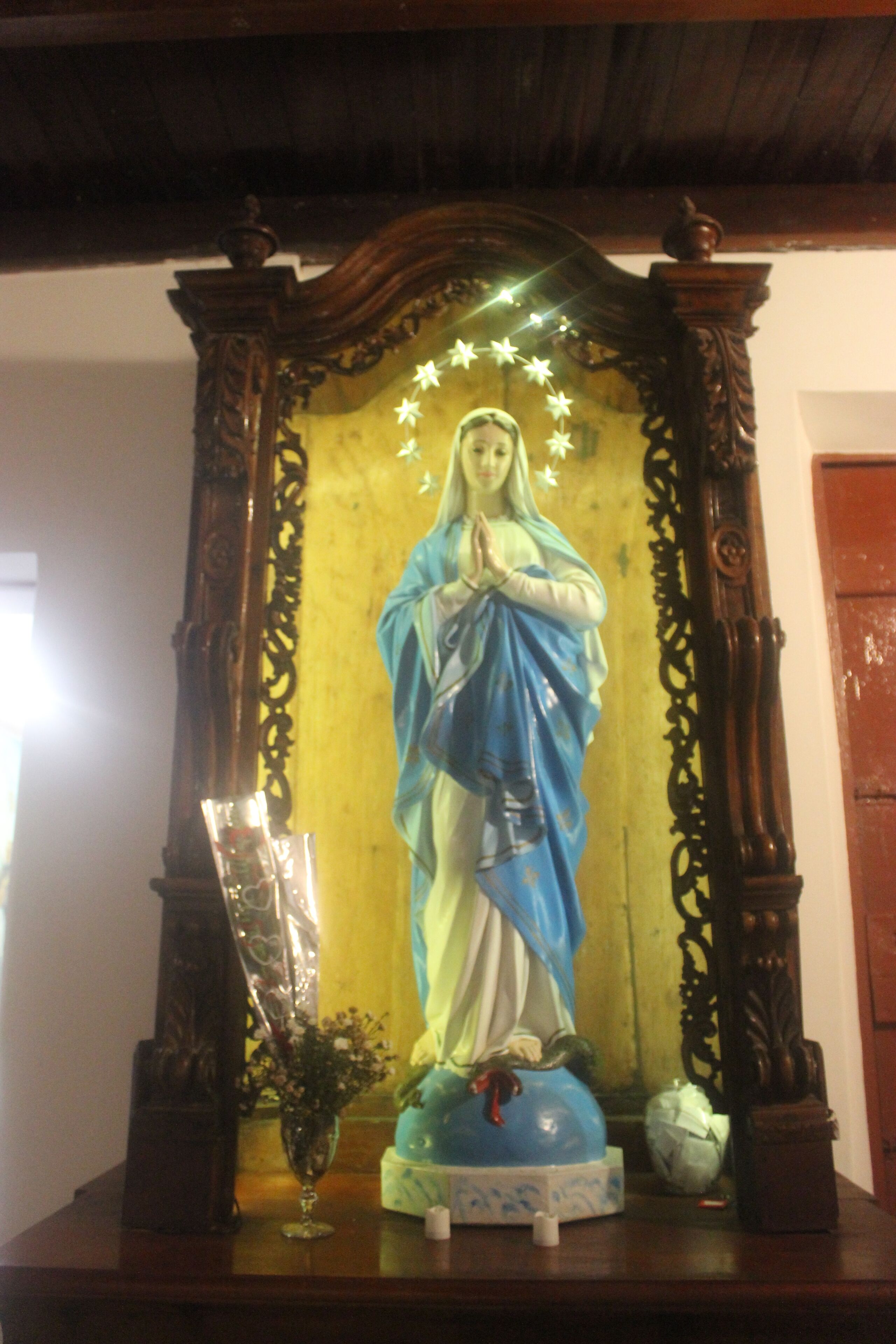Een standbeeld van een religieuze figuur met een halo, omringd door versierde houtsnijwerken en een vaas met bloemen.