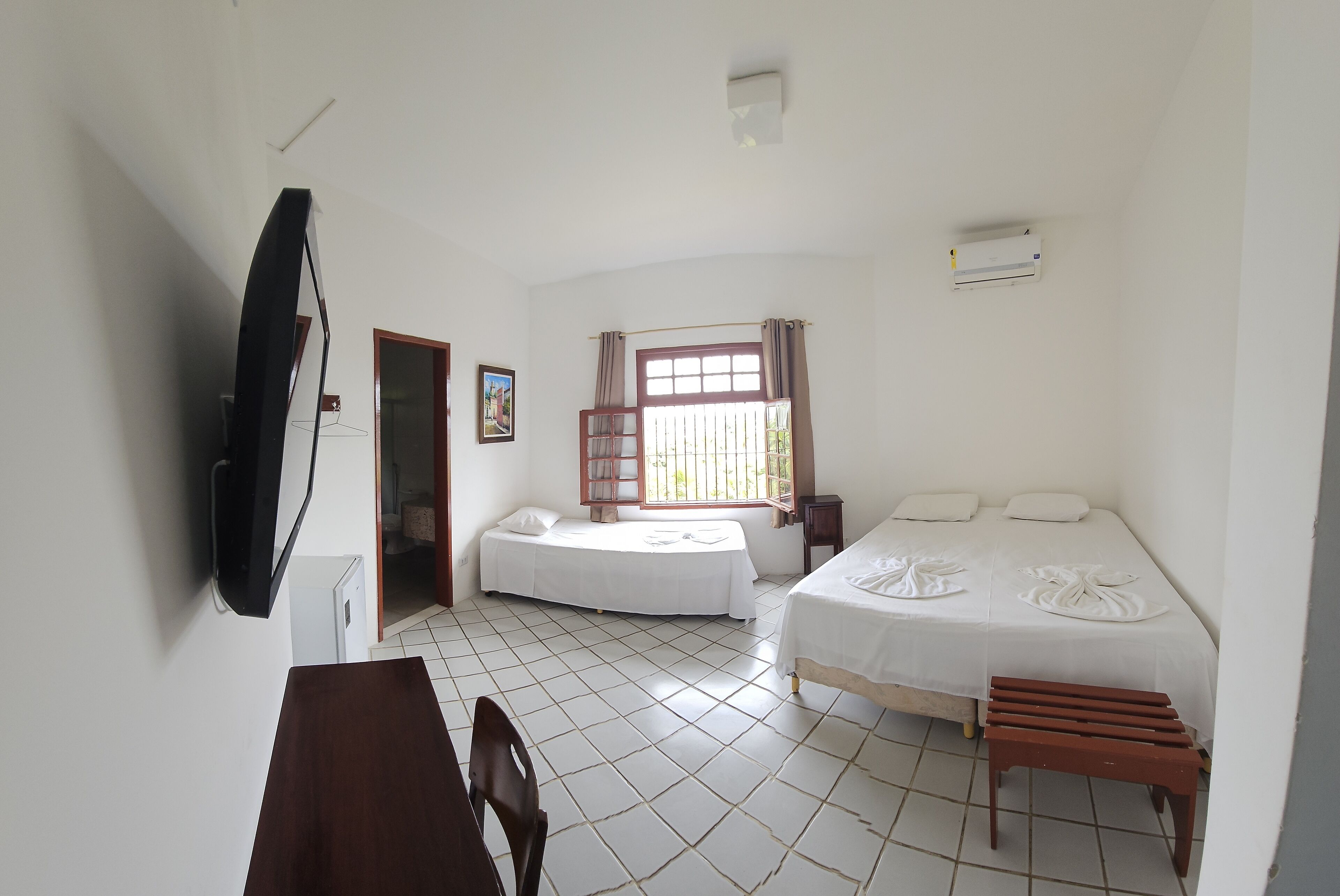 A room with two beds, a TV mounted on the wall, a window with curtains, and a tiled floor.