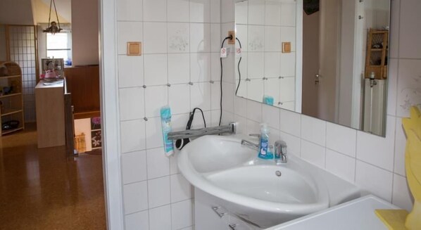 Ein Badezimmer mit einem weißen Waschbecken, einem Spiegel und gefliesten Wänden. Ein Holzschrank und ein Regal mit Gegenständen befinden sich links.