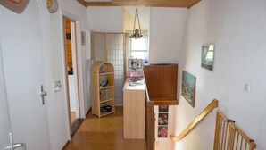 Un pasillo estrecho con una escalera de madera que lleva a una plataforma. Se ve en el fondo una estantería de madera con diversos objetos.