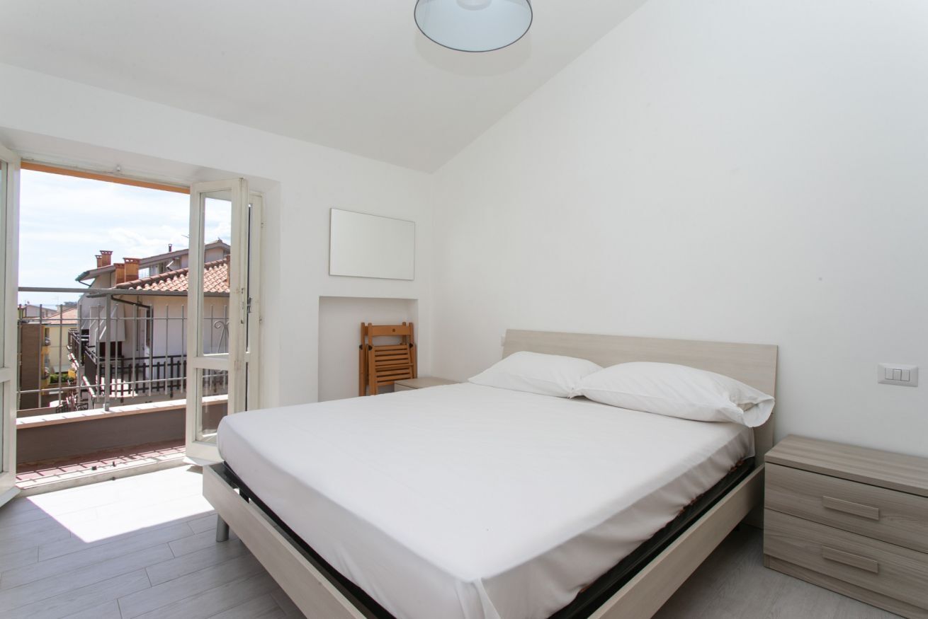 A bedroom with a bed, a nightstand, a chair, and a balcony with a view of buildings.