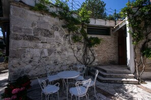 Un muro de piedra con una mesa y sillas de metal, un árbol pequeño y una puerta.