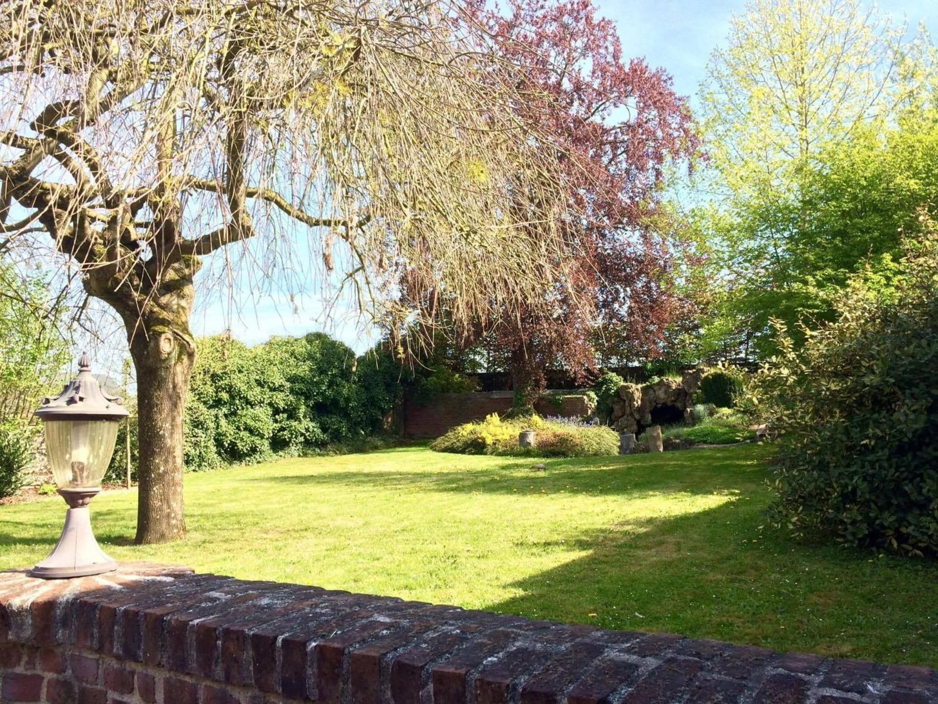 A well-maintained garden with a variety of trees, a brick wall, and a lamp post.