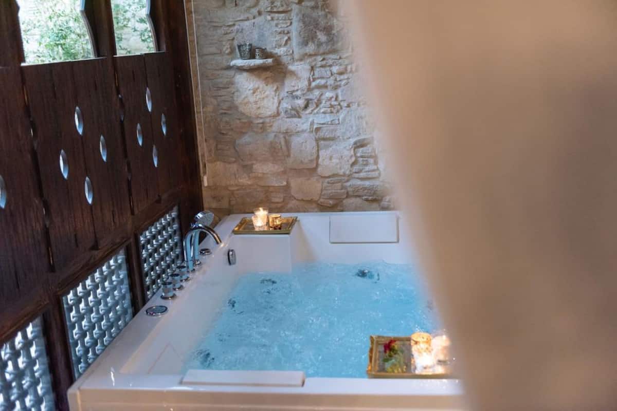 Ein Badezimmer mit Steinwänden, einer Badewanne, einer Holztür mit runden Ausschnitten und einem Regal mit Kerzen.