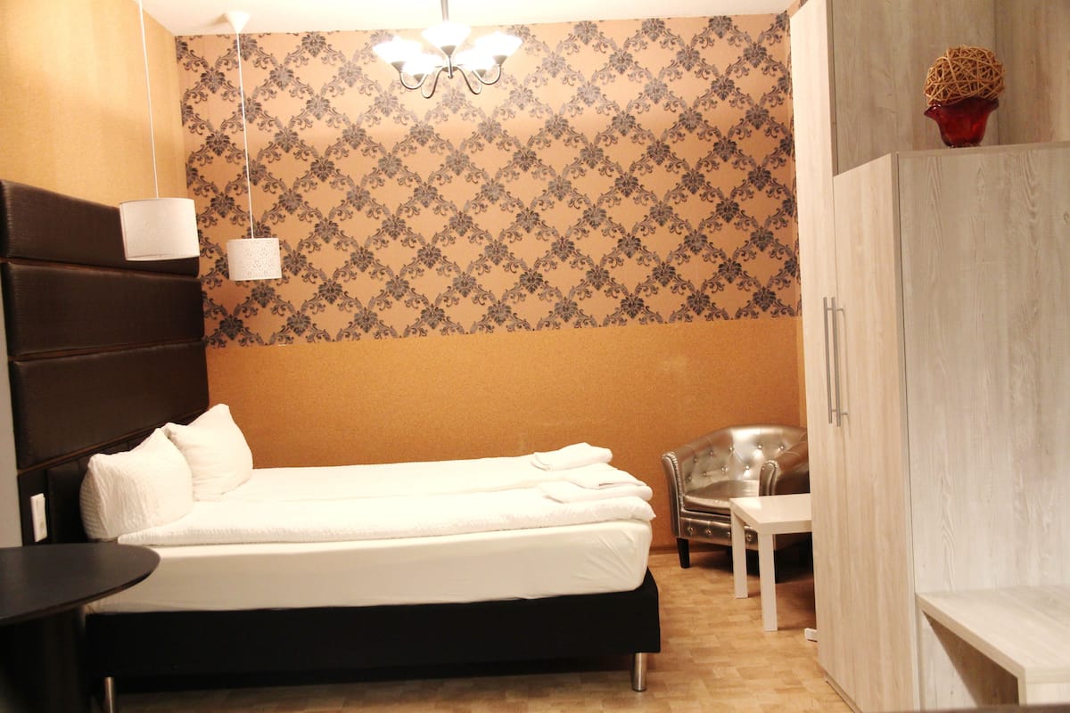 Una habitación de hotel con cama, una mesita, una silla, un armario y un papel tapiz estampado.
