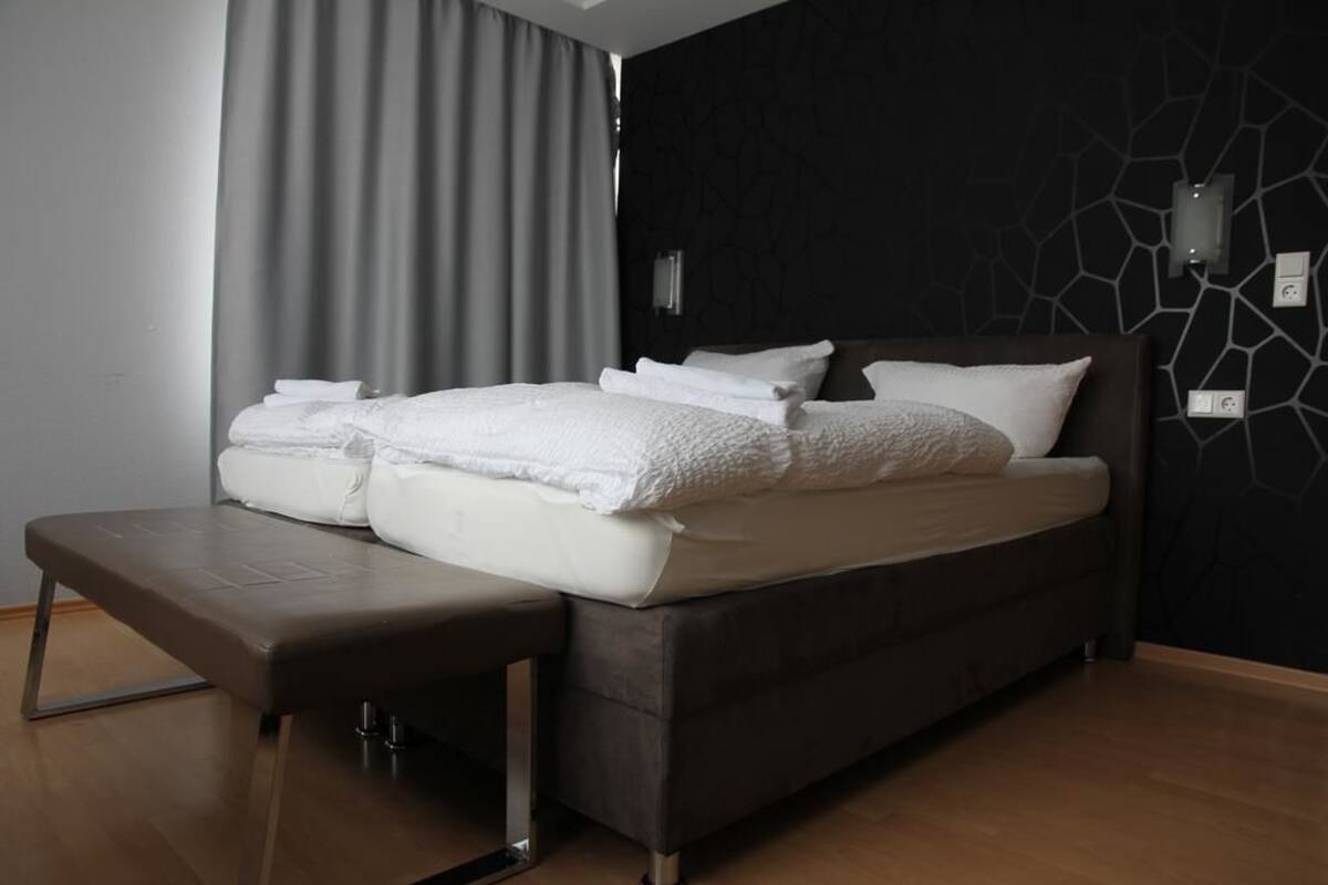 Un dormitorio moderno con una cama grande, un banco y una pared oscura con un diseño geométrico.