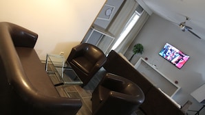 A modern living room with a sofa, armchairs, a glass coffee table, a flat-screen TV mounted on the wall, a ceiling fan, and a clock on the wall. - Apartment Caplex (Oldenburg)