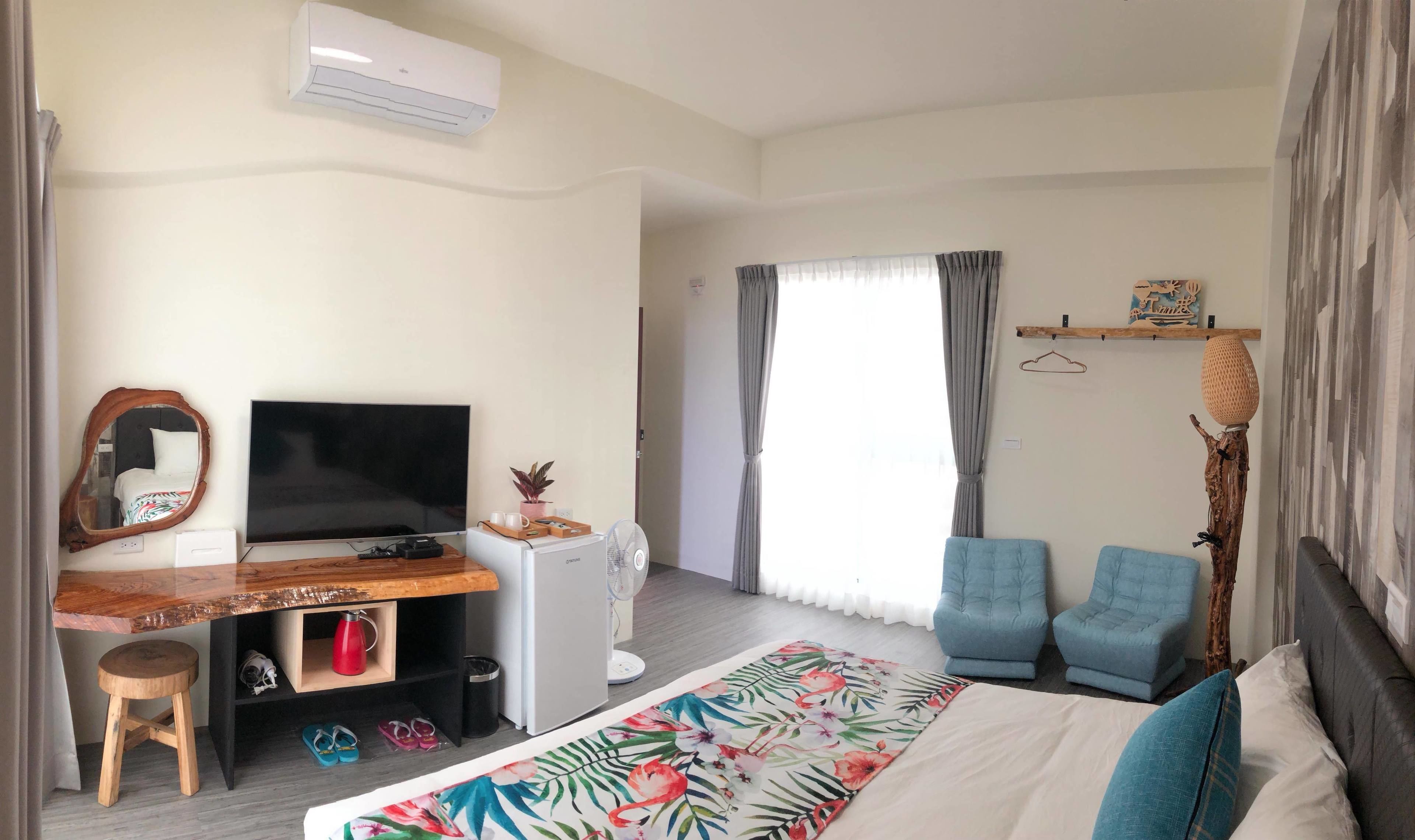 A hotel room with a bed, a TV, a desk with a computer, two chairs, a window with curtains, and a wall-mounted air conditioner.