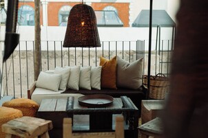 A cozy outdoor seating area with a wicker hanging light fixture, cushioned seating with pillows, and a wicker basket.