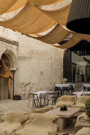 Una zona d'exterior per dinar amb taules i cadires, un mur de pedra rústic i una finestra gran.