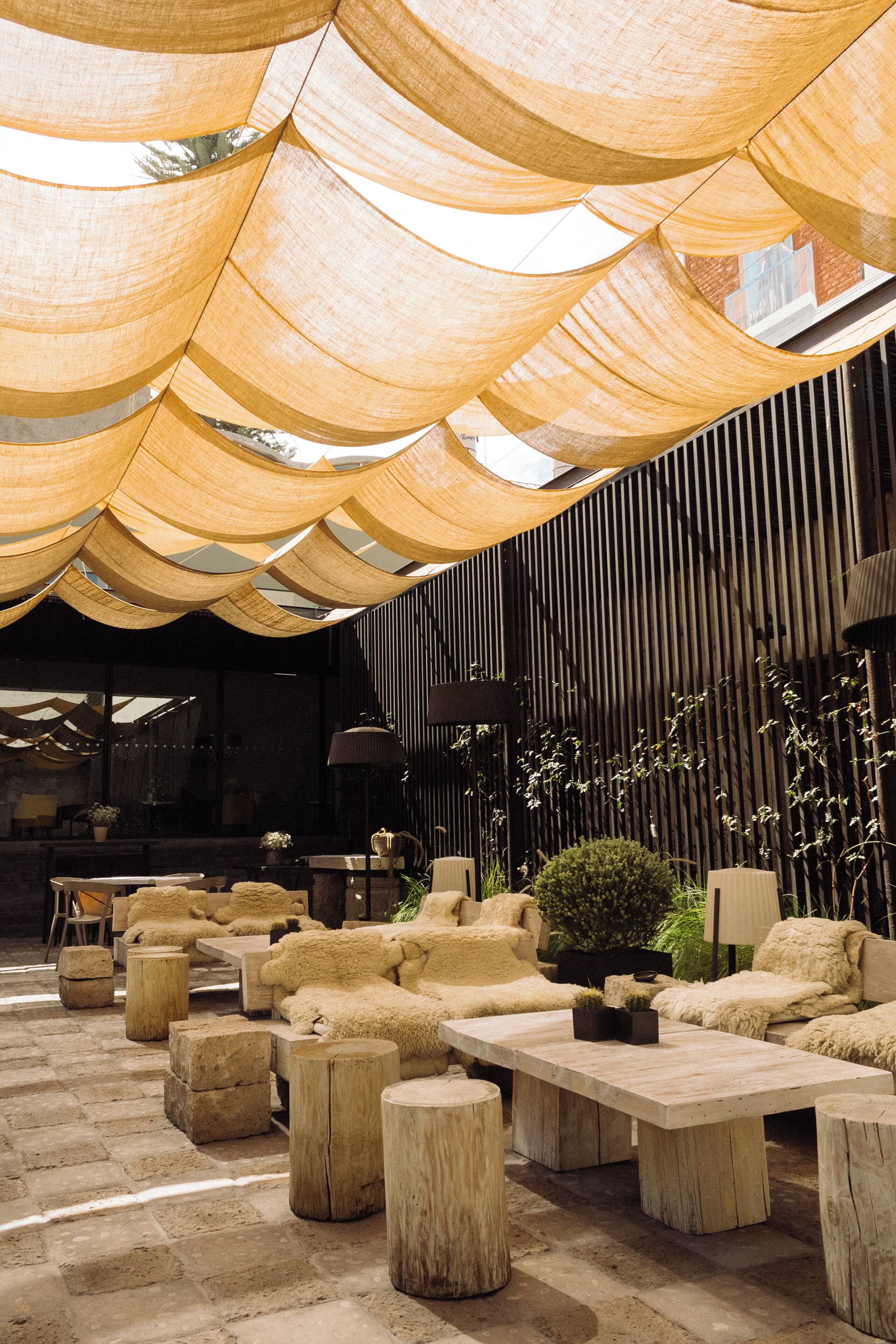 A modern outdoor seating area with wooden tables and chairs, a stone floor, and a canopy overhead.
