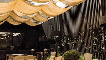 A modern outdoor seating area with wooden tables and chairs, a stone floor, and a canopy overhead.