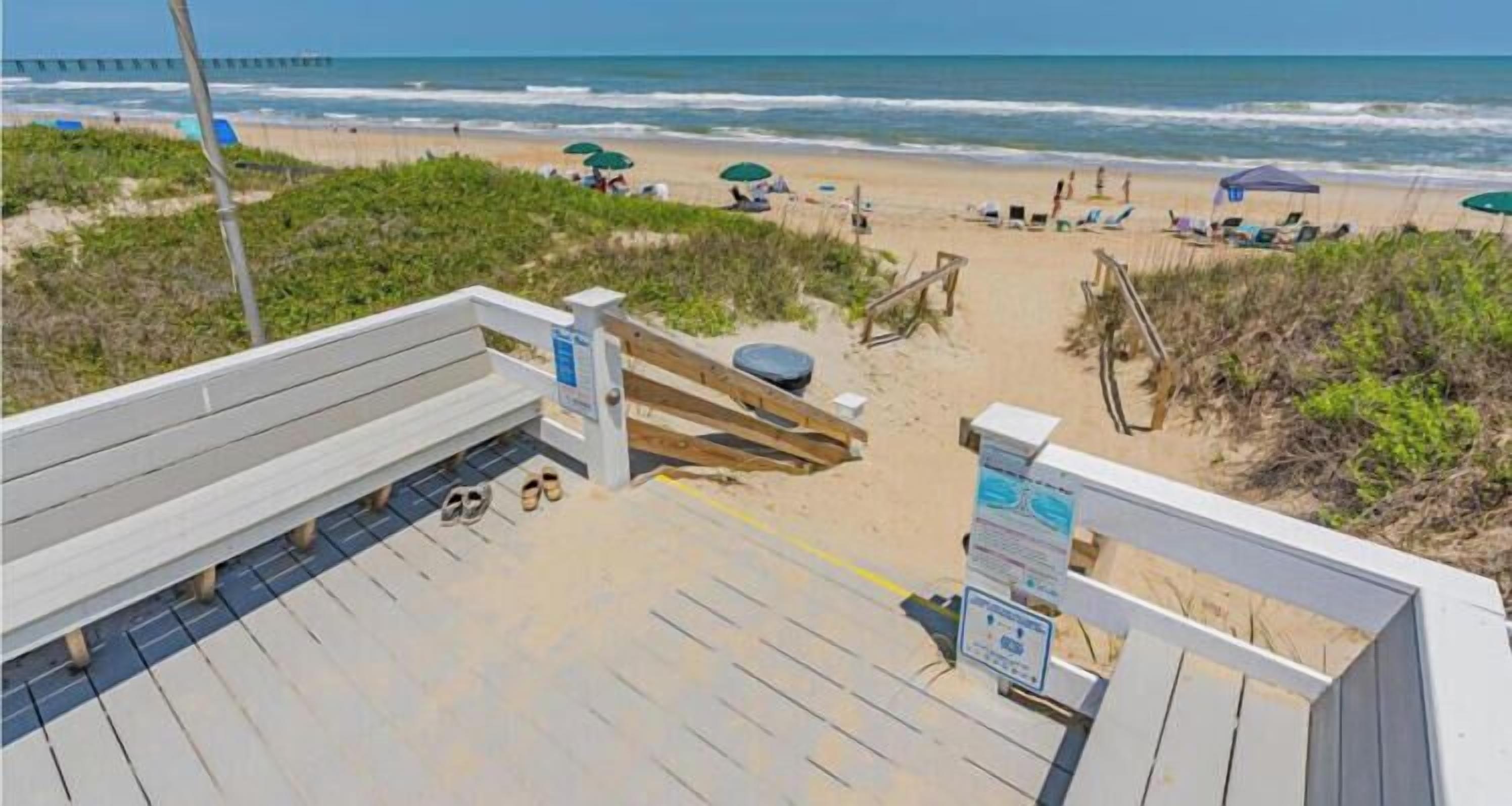 A beachside walkway with a wooden bench, a lifeguard stand, and beach umbrellas.