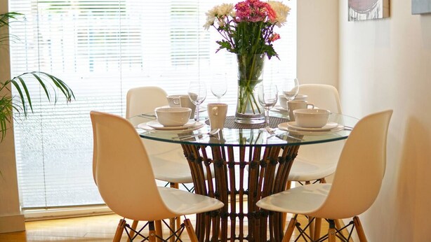 A dining area with a glass table surrounded by white chairs, a vase with flowers, and a window with blinds. - Superior Canary Wharf  O2 Arena 2Bed Apartment (Greater London)