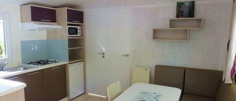 Cocina compacta con estufa a gas, microondas y un refrigerador pequeño. Hay una zona de comedor con una mesa y sillas, y un sofá.