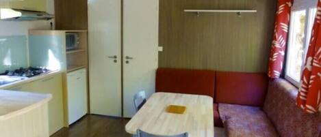 A compact kitchen with a stove, a refrigerator, and a small sink. There is a dining table with two chairs and a bench with a cushion.