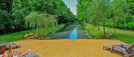 A river with trees on both sides, a sandy area with lounge chairs, and a person sitting on one of the chairs.