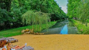 Un río flanqueado por árboles, una zona arenosa con sillas de descanso y una persona sentada en una de ellas.